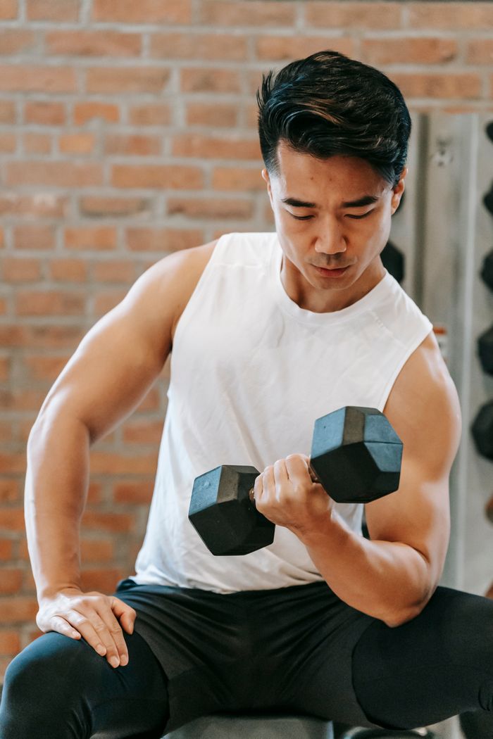 From below of focused muscular male athlete doing bicep curls with dumbbell against brick wall in contemporary gym